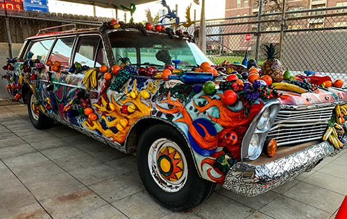 An old wagoneer-styled car is completely covered in vibrantly painted fake fruits and bright flames.
